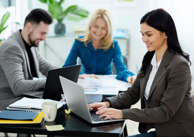 Business directory image of three professionals collaborating and smiling while working on laptops around an office table